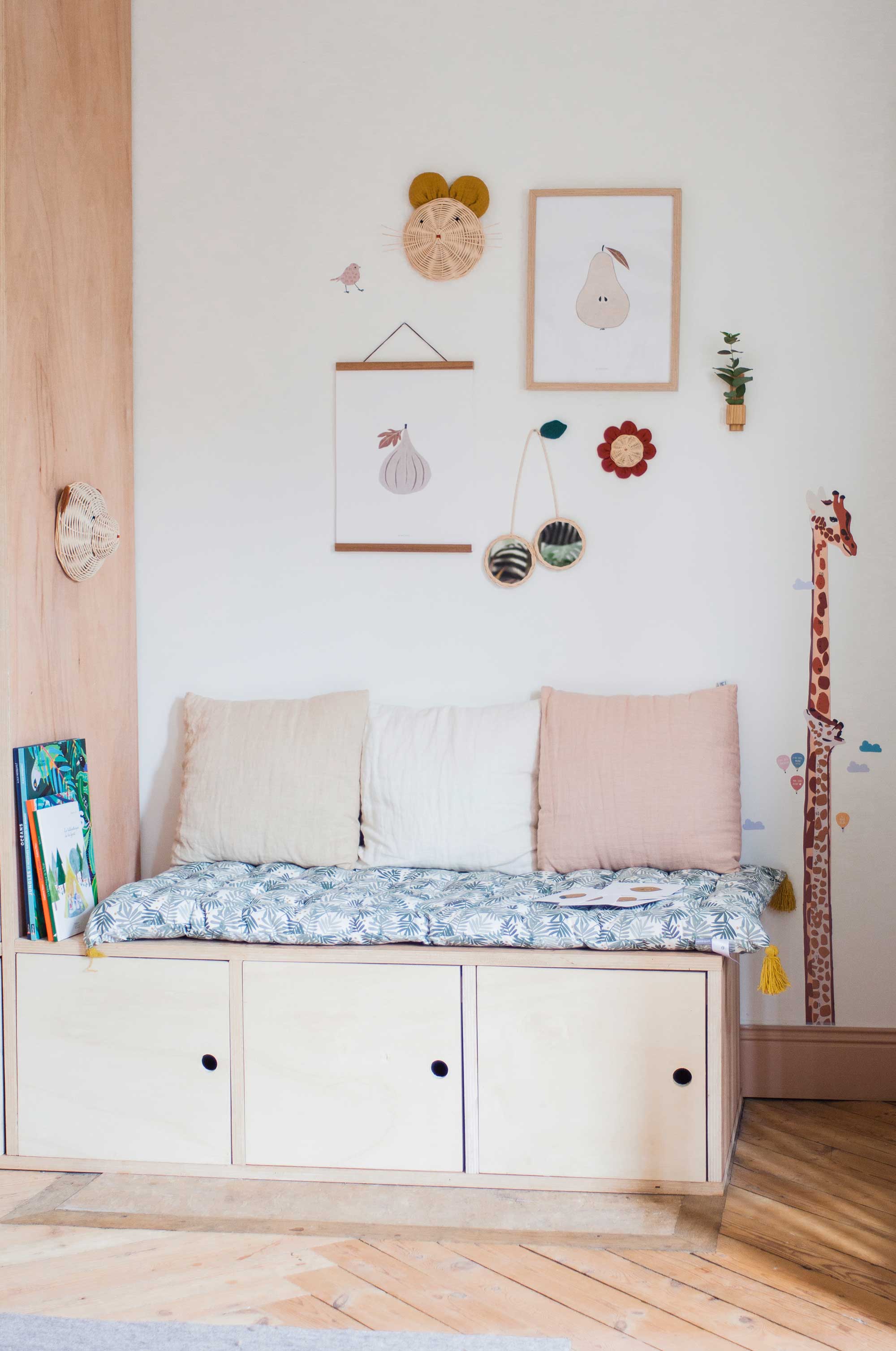 un coin est aménagé avec une banquette et des coussins. Au dessus, sur le mur, sont accrochés des objets de décoration en rotin. Il y a un trophée souris, un trophée ours,un miroir cerise et une fleur. Il y a également des illustrations encadrées.