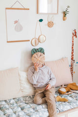 Un enfant est assis sur une banquette et cache son visage derrière un trophées en rotin en forme de souris. Au dessus de lui on voit d'autres objets en rotin accrochés au mur : un miroir cerise et un trophée lapin.